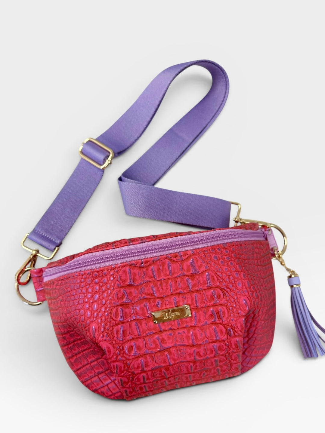 Pink croc-embossed belt bag with purple zipper, lavender tassel, and wide lavender crossbody strap