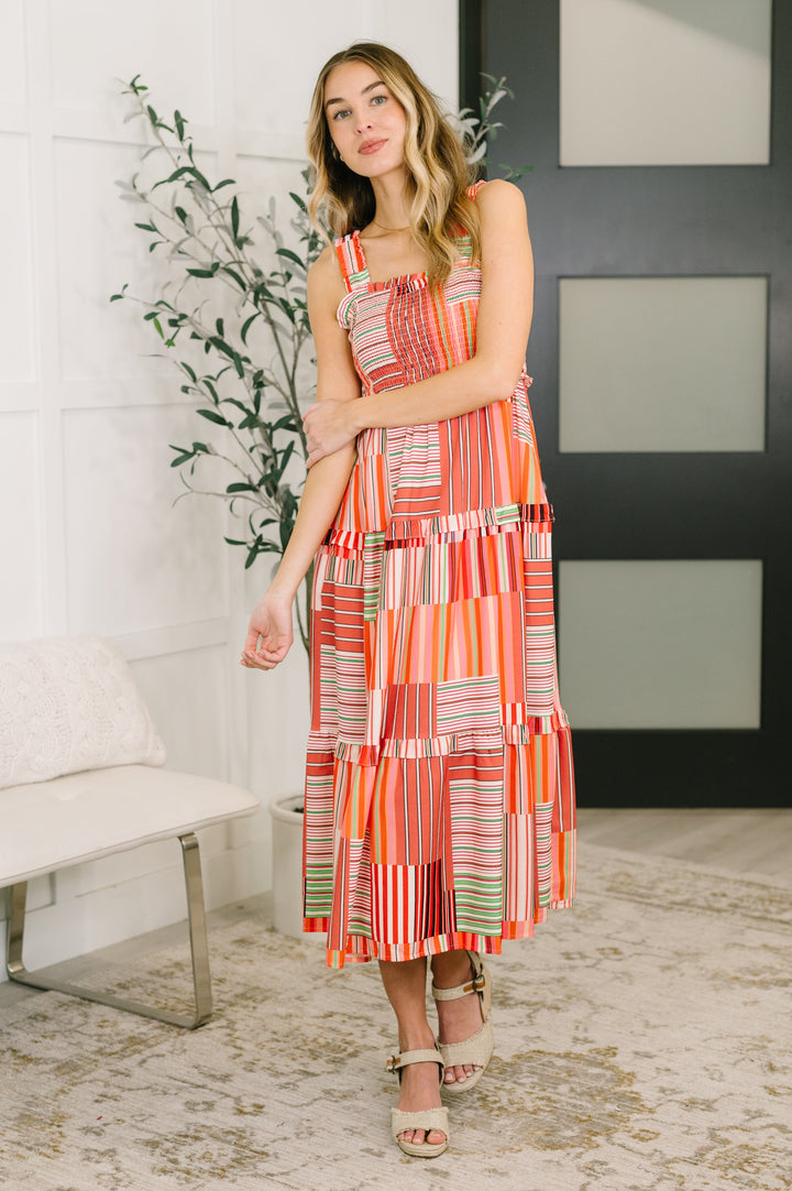 Three-quarter front view of a woman in the coral patchwork stripe smocked midi dress with tan wedge sandals.
