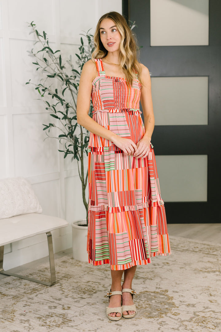 Full-length front view of a woman in a coral multicolor patchwork stripe smocked bodice midi dress with tiered skirt, paired with tan wedge sandals.