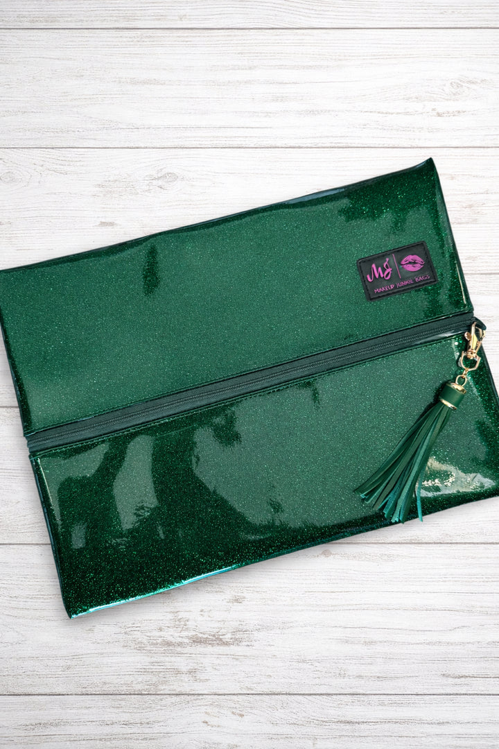 Makeup Junkie Bags emerald green glitter foldover zip makeup bag with green tassel zipper pull and branded patch, front view on white wood background