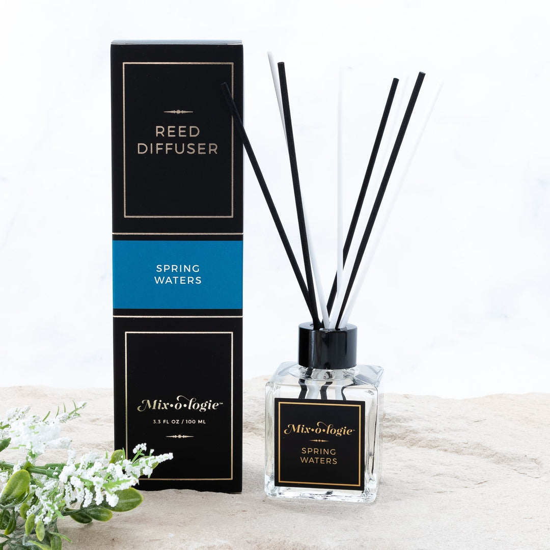 Mixologie Reed Diffuser in Spring Waters, 3.3 fl oz, clear square glass bottle with black cap and black label with blue accent band, with matching black and blue box, styled with white florals