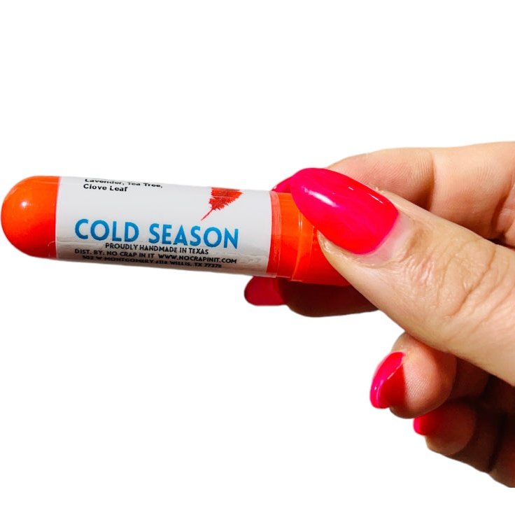 Hand with hot pink nails holding a No Crap In It Cold Season essential inhaler with orange cap, made with lavender, tea tree, and clove leaf, on a white background.