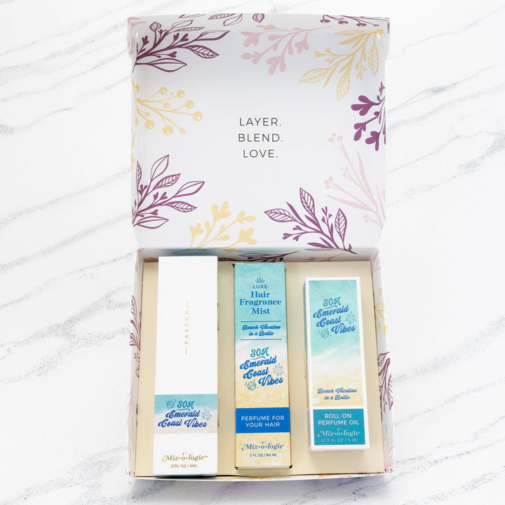 Mixologie Party of Three Gift Set in 30A Emerald Coast Vibes open box showing parfum spray, Luxe Hair Fragrance Mist, and roll-on perfume oil in teal and sand packaging, top view on marble background