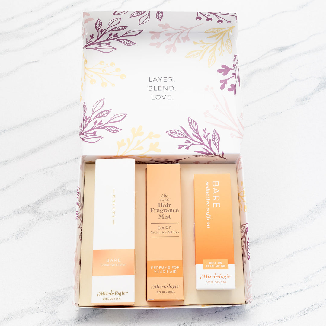 Mixologie Party of Three Gift Set in Bare Seductive Saffron open box showing parfum spray, Luxe Hair Fragrance Mist, and roll-on perfume oil in peach and orange packaging, top view on marble background