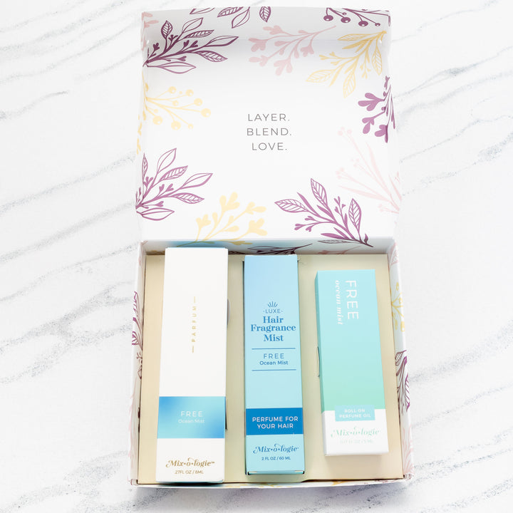 Mixologie Party of Three Gift Set in Free Ocean Mist open box showing parfum spray, Luxe Hair Fragrance Mist, and roll-on perfume oil in light blue packaging, top view on marble background