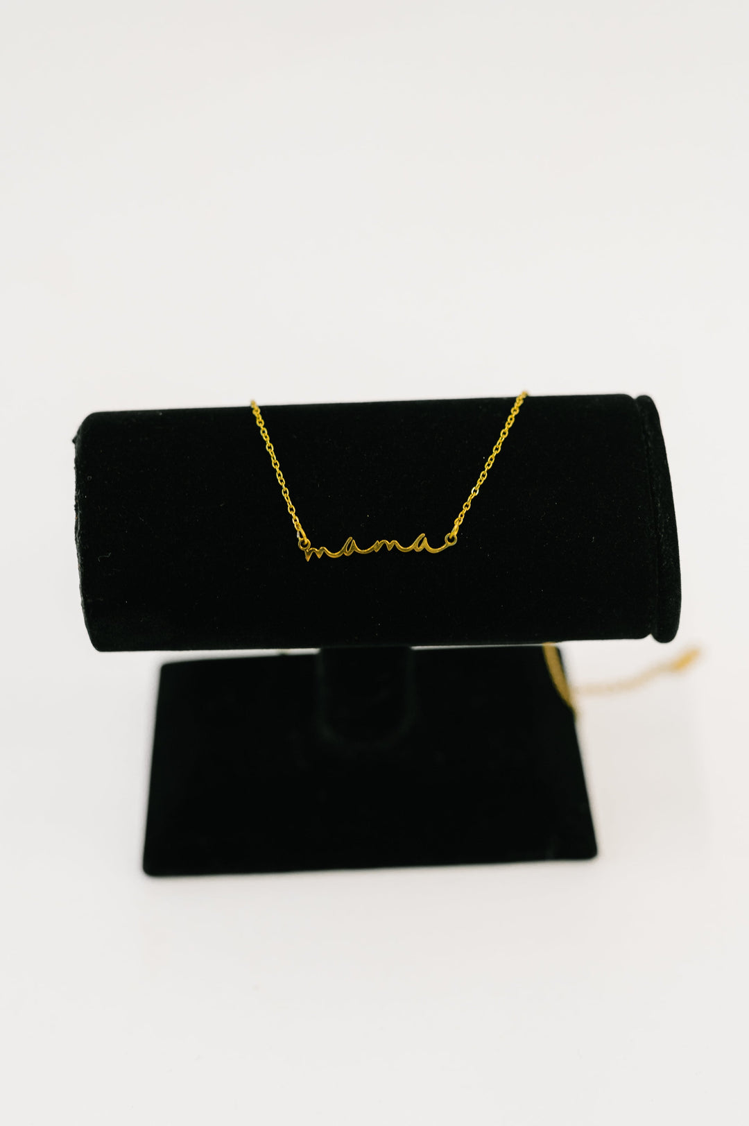 Gold "mama" script necklace displayed on a black jewelry stand against a white background