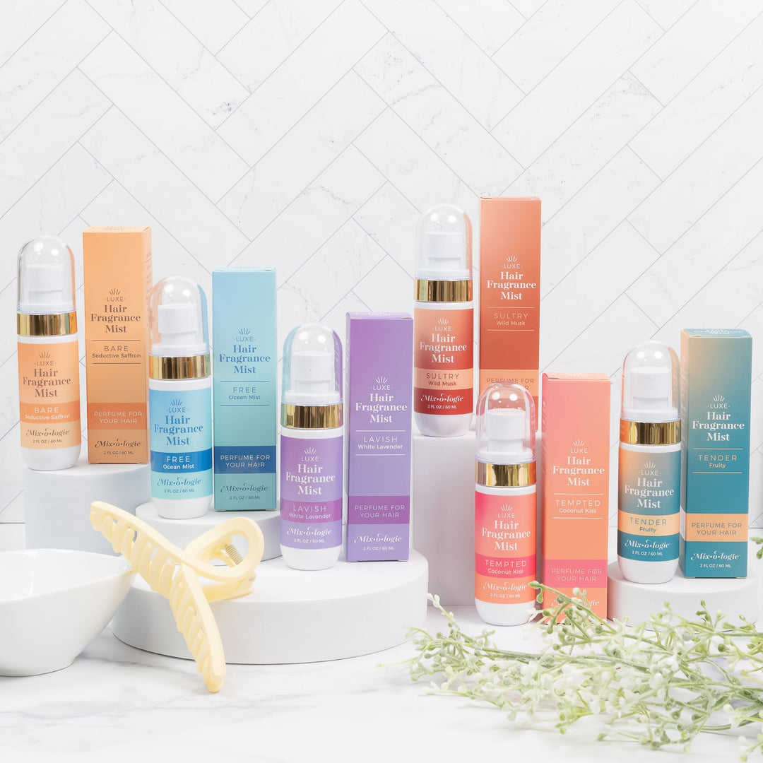 Mixologie Luxe Hair Fragrance Mist full collection in six scents including Bare, Free, Lavish, Sultry, Tempted, and Tender displayed with bottles and boxes on white risers with hair clip and greenery, lifestyle shot
