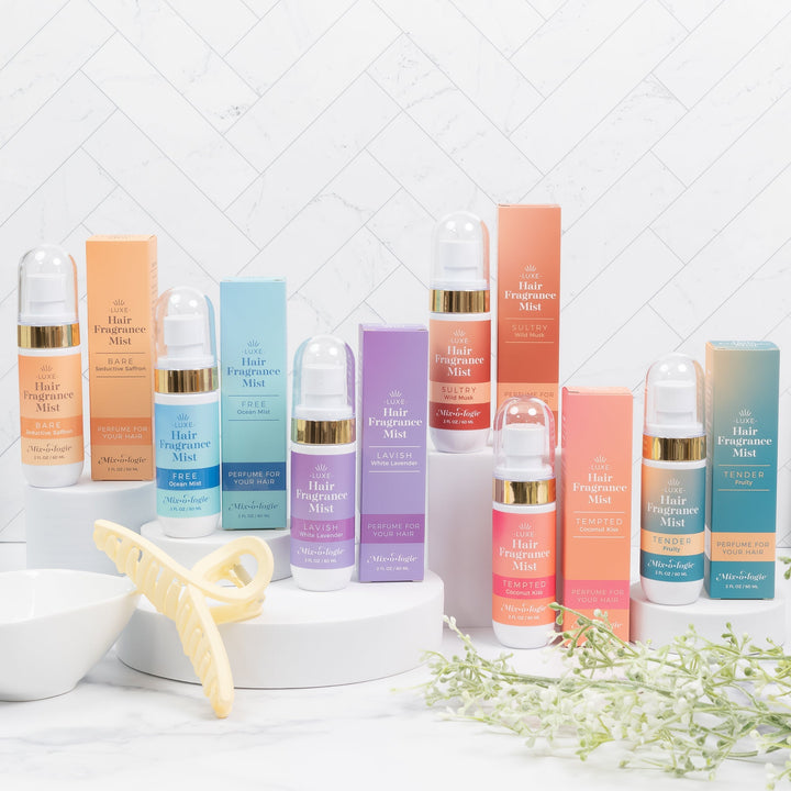 Mixologie Luxe Hair Fragrance Mist full collection in six scents including Bare, Free, Lavish, Sultry, Tempted, and Tender displayed with bottles and boxes on white risers with hair clip and greenery, lifestyle shot