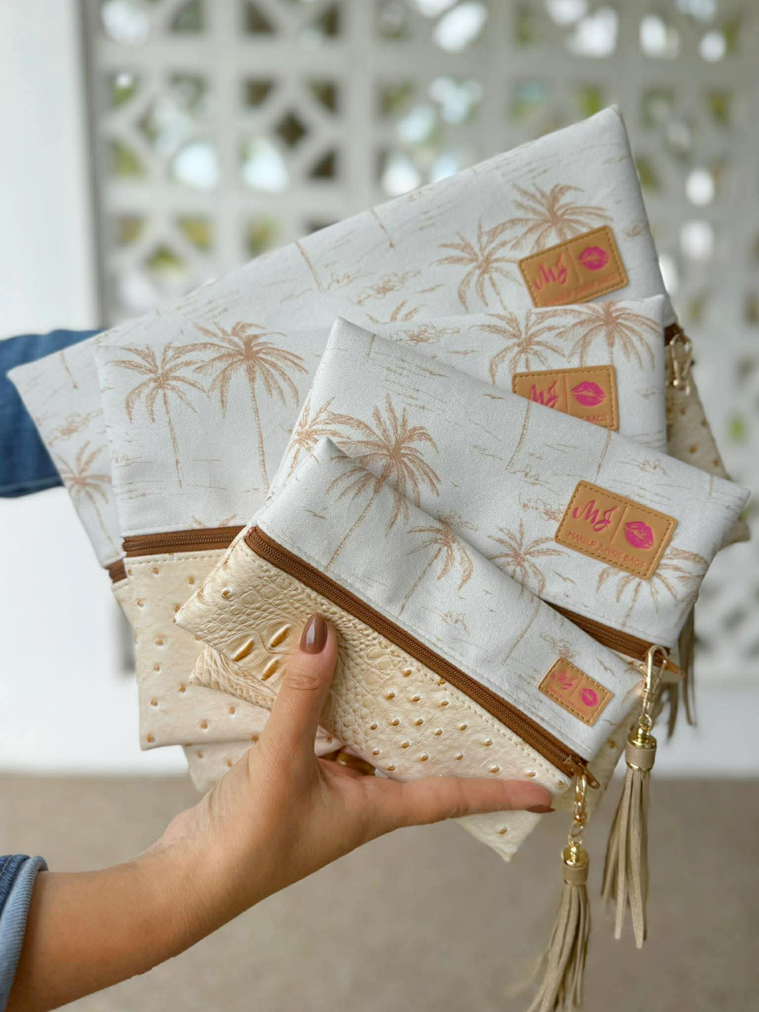 Hand holding a fanned stack of four white and gold palm tree print zip pouches in graduated sizes, with the front pouch featuring a cream ostrich-embossed panel with gold studs, tan zipper, and gold tassel pull, against a white lattice background.