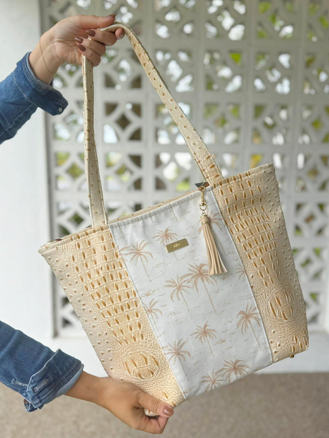 Hand holding a white and gold palm tree print tote bag with cream croc-embossed side panels, dual tan leather handles, a gold embossed logo at the base, and a tan tassel zipper pull, against a white lattice background.