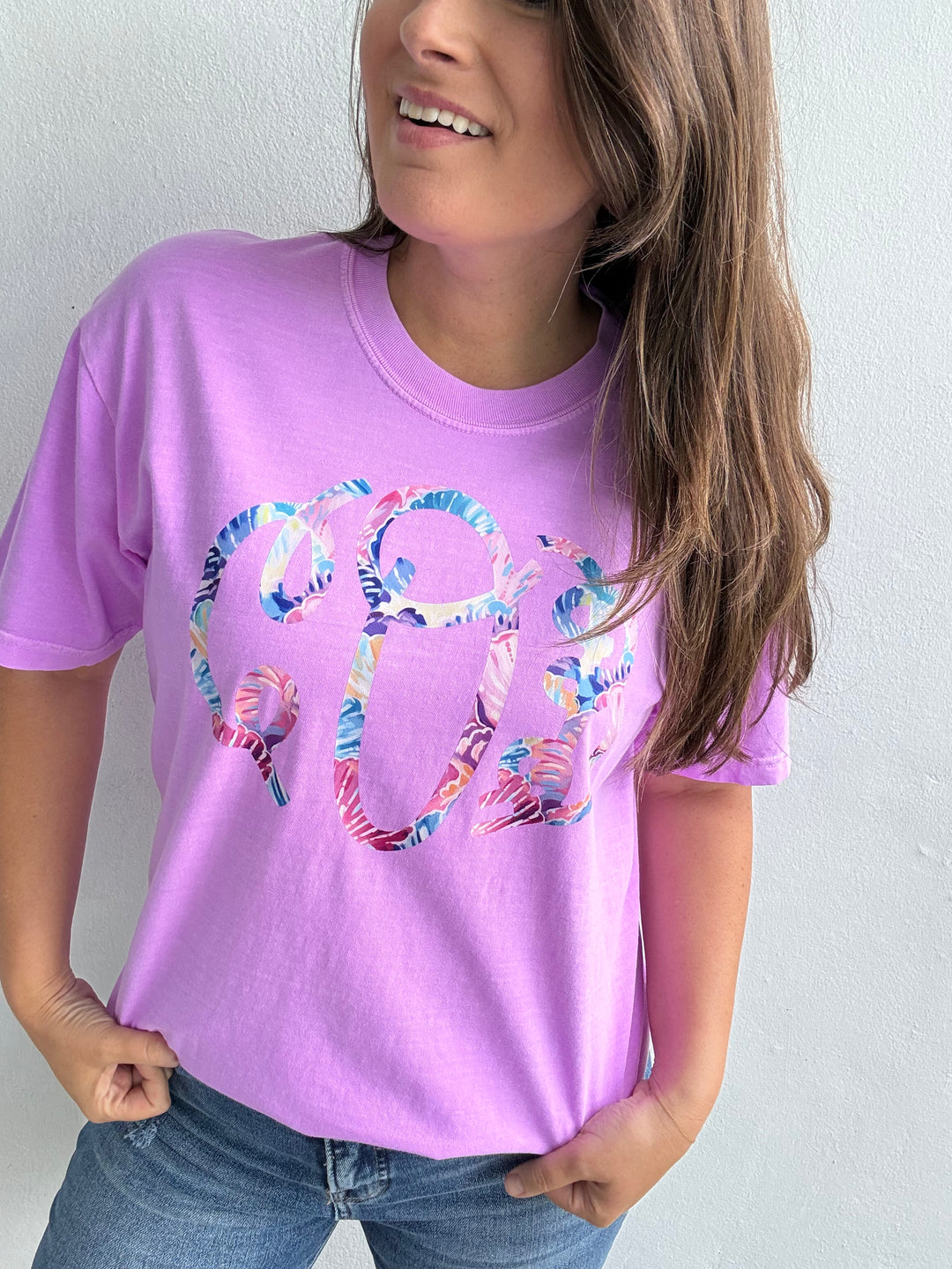 Woman wearing a lavender Comfort Colors t-shirt with a large colorful floral monogram graphic, posed against a white wall