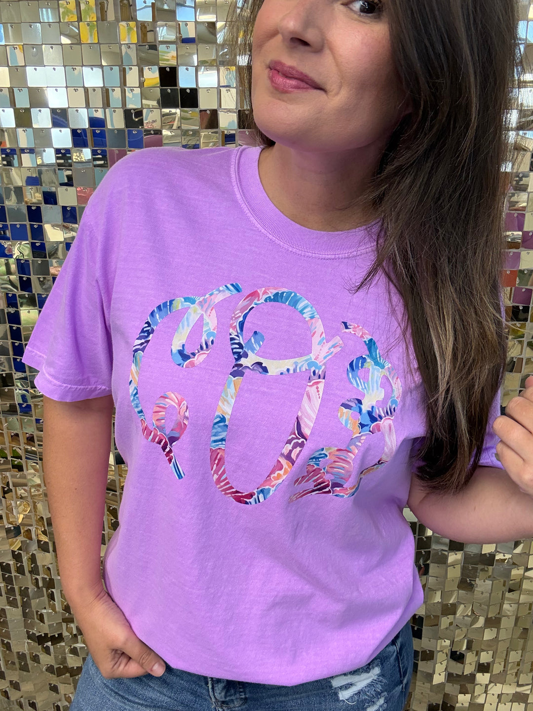 Woman wearing a lavender Comfort Colors t-shirt with a large colorful floral monogram graphic, posed in front of a sequin backdrop
