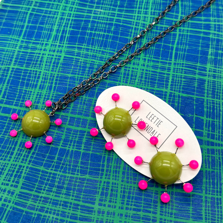 Olive green pendant with hot pink beads radiating around the center, reminiscent of the Sputnik satellite. Pendant hangs on an adjustable chain, matching earrings are shown alongside. Jewelry is arranged on a blue and green plaid background.