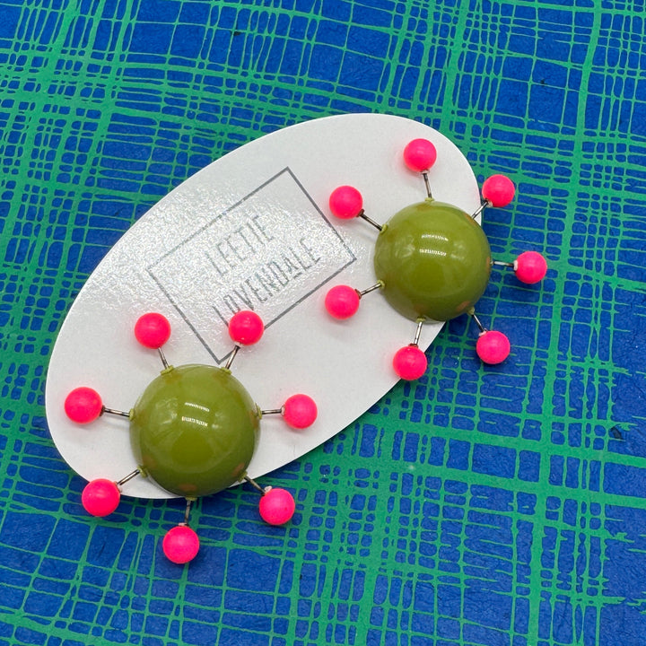 OlIve green earrings with hot pink beads radiating from center with posts on a blue and green plaid background.