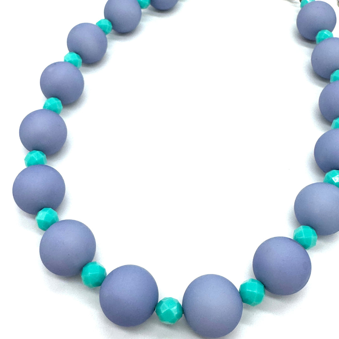 Closeup of necklace featuring chunky periwinkle lucite beads and smaller faceted turquoise spacers on a white background.