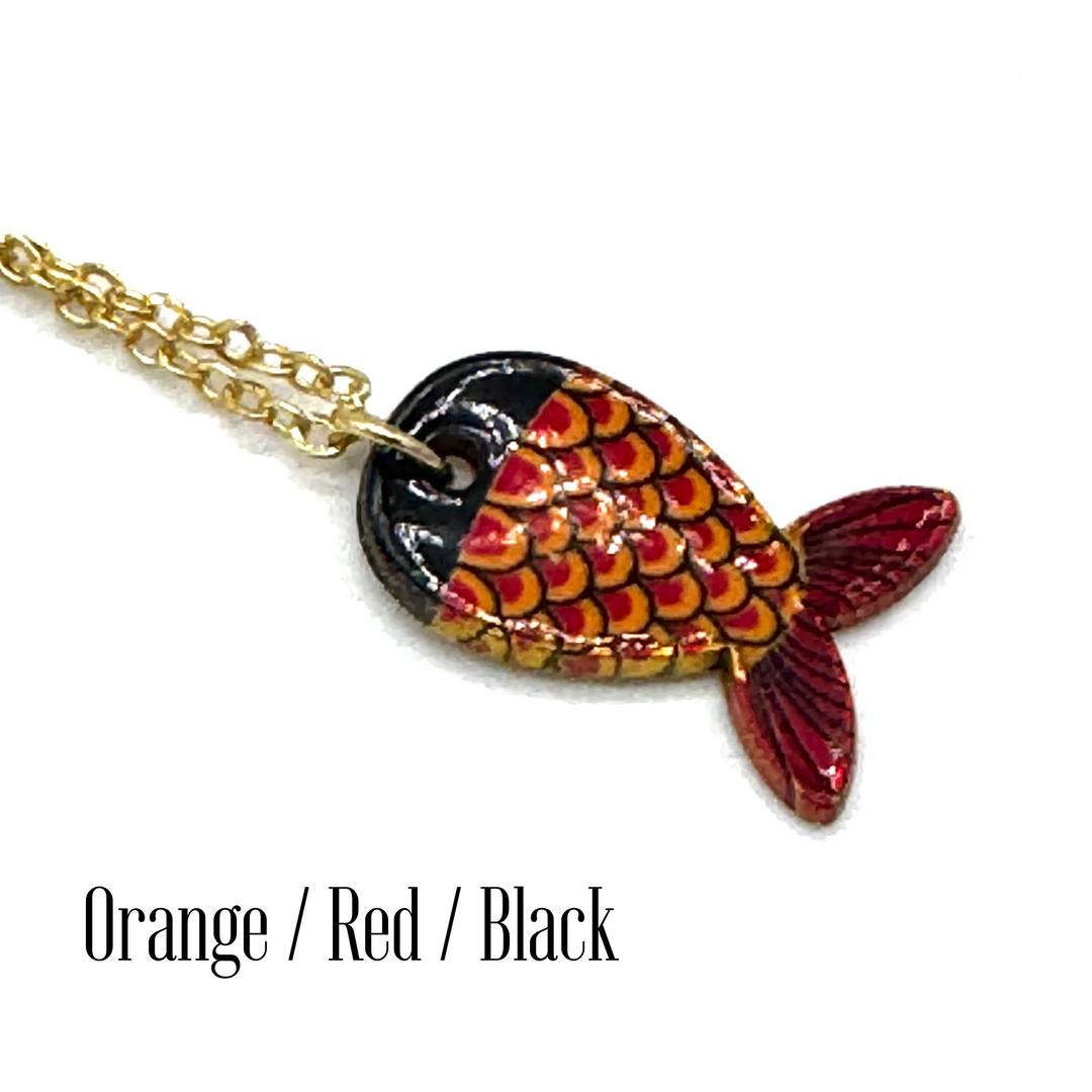 A close-up of a fish-shaped pendant necklace on a gold chain against a white background, with the text 'Orange / Red / Black' below. The pendant features a glossy black top half, orange and red scalloped scales, and a red tail.