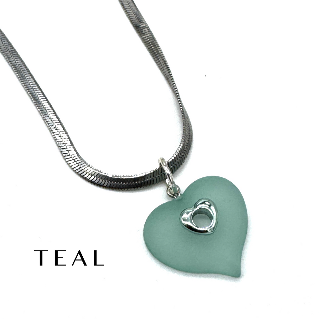 A teal heart-shaped pendant necklace with a silver snake chain and a small silver heart detail, shown on a white background with the word &