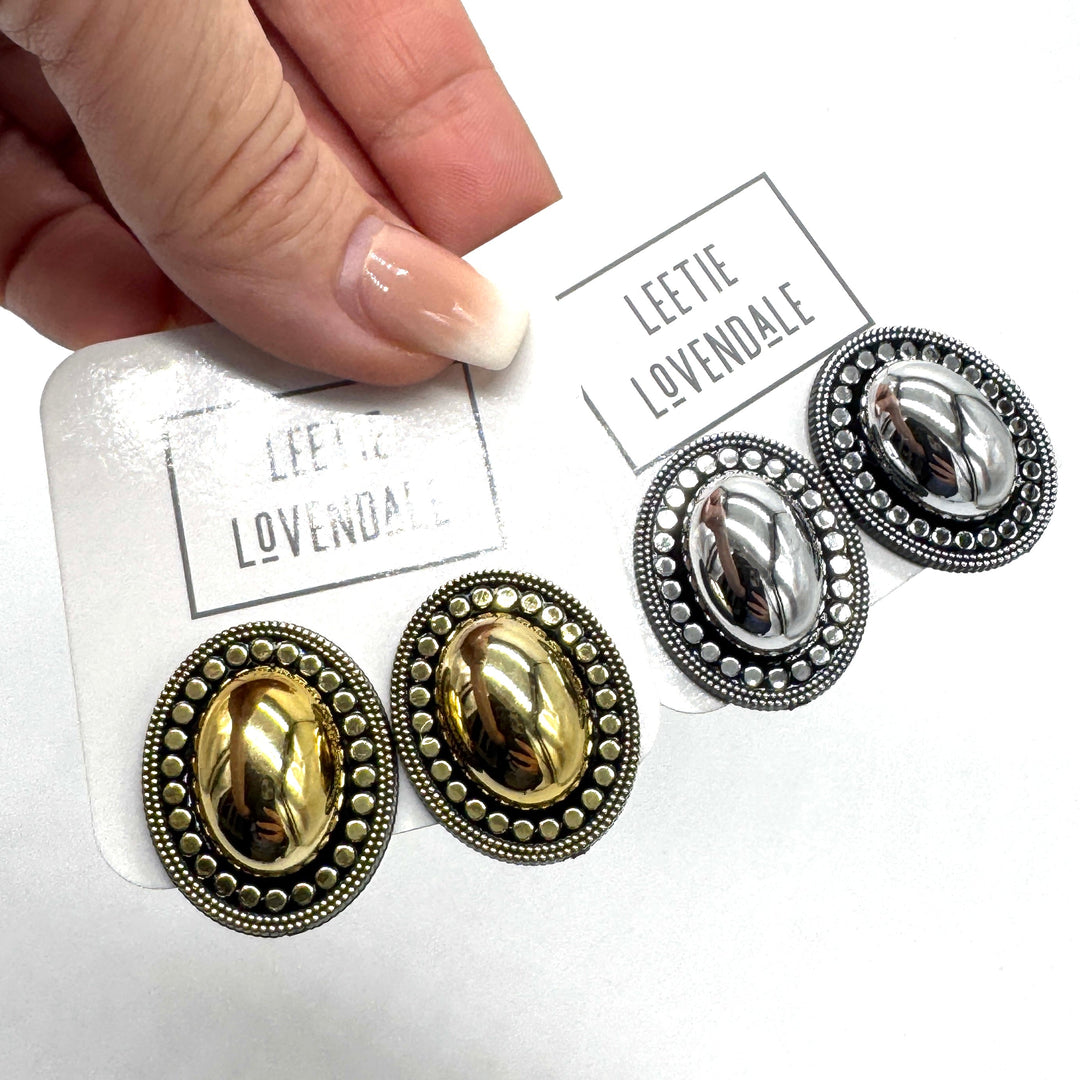 A close-up of a hand holding a white card displaying two pairs of oval stud earrings on a white background. The pairs are gold-toned and silver-toned with ornate beaded borders and smooth oval centers.