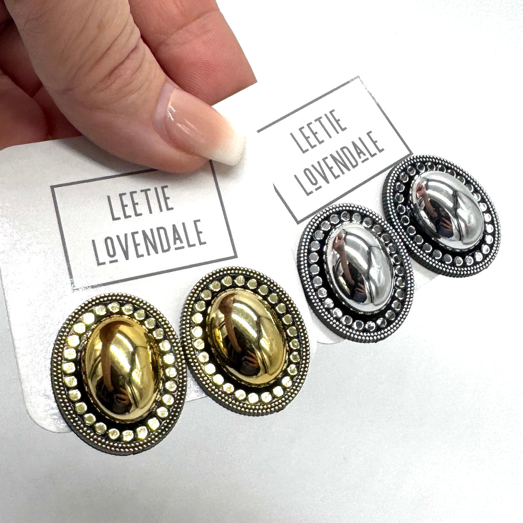 A close-up of a hand holding a white card printed with the brand name 'LEETIE LOVENDALE' above a pair of oval stud earrings. The card also shows a second pair of matching earrings.