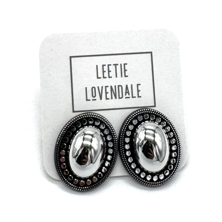 A pair of oval stud earrings with shiny silver-toned centers and dark textured outer borders, displayed on a white branded backing card that reads LEETIE LOVENDALE.
