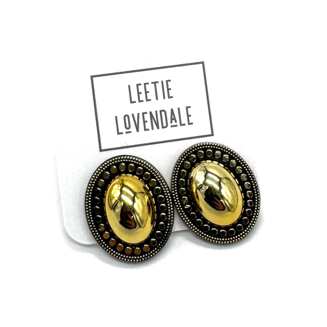 A pair of oval stud earrings with a shiny gold center and a dark bronze textured border, displayed on a white backing card with the brand name 'LEETIE LOVENDALE' printed above them.