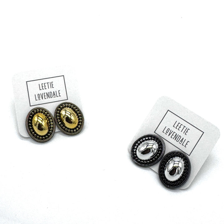 Two pairs of oval stud earrings on white display cards. One pair features gold metal with a textured border, and the other pair features silver metal with a textured border.