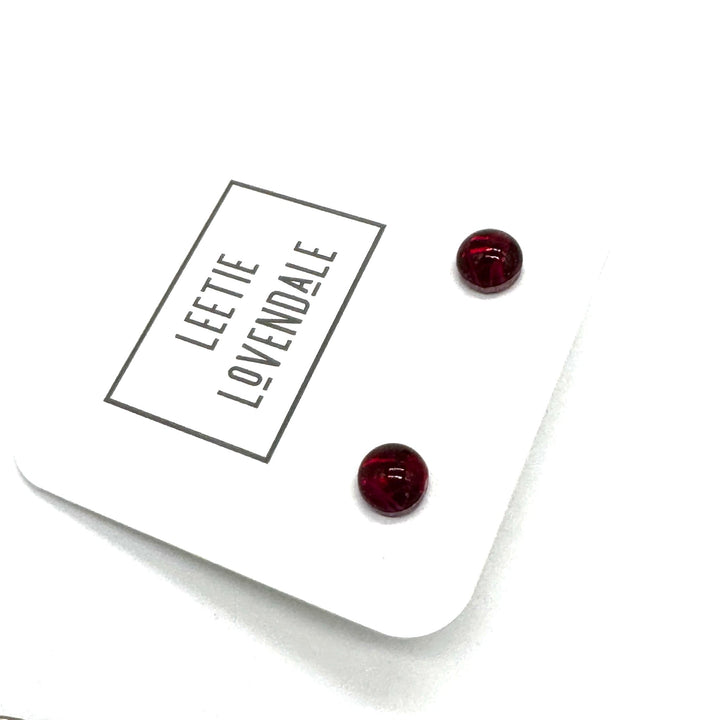 A pair of small round stud earrings in deep red resin, displayed on a white branded card that reads &