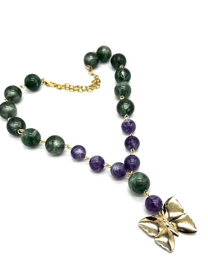A beaded necklace featuring dark green and purple lucite beads connected by small gold links, with a gold butterfly pendant and gold chain clasp, shown on a plain white background.