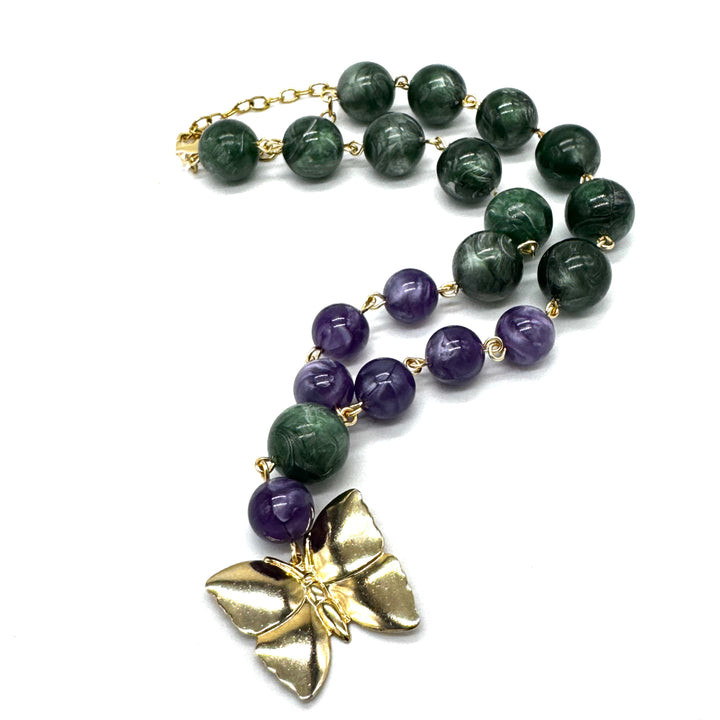 A beaded necklace featuring dark green and purple lucite beads separated by small gold links, with a large textured gold butterfly pendant at the end, displayed on a clean white background.