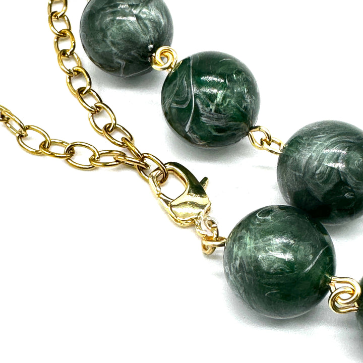 A close-up of a beaded necklace featuring polished dark green lucite beads with white marbling, connected by a gold-tone chain and clasp, on a white background.