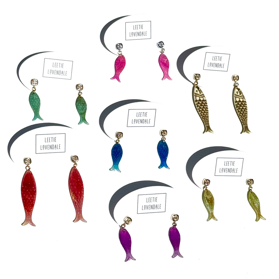 Six pairs of fish-shaped dangling earrings arranged on a white background, each with a small brand label reading 'LEETHE LOVENDALE'. The earrings come in green, blue, red, purple, and gold-tone finishes.