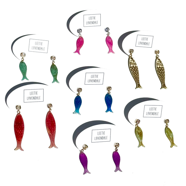 Six pairs of fish-shaped dangling earrings arranged on a white background, each with a small brand label reading 'LEETHE LOVENDALE'. The earrings come in green, blue, red, purple, and gold-tone finishes.