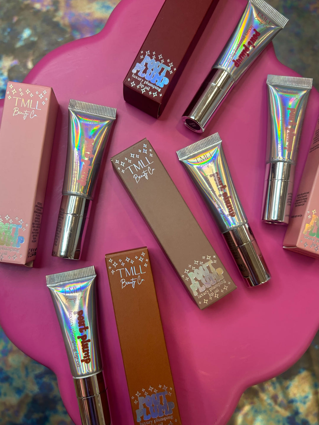 TMLL Beauty Co Pout Plump luxury plumping lip oil holographic silver tubes and boxes in multiple shades arranged on pink surface
