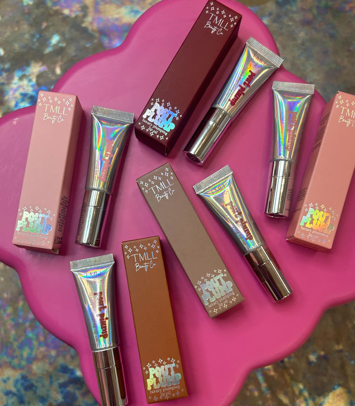 TMLL Beauty Co Pout Plump luxury plumping lip oil holographic silver tubes and boxes in pink and brown shades arranged on pink surface