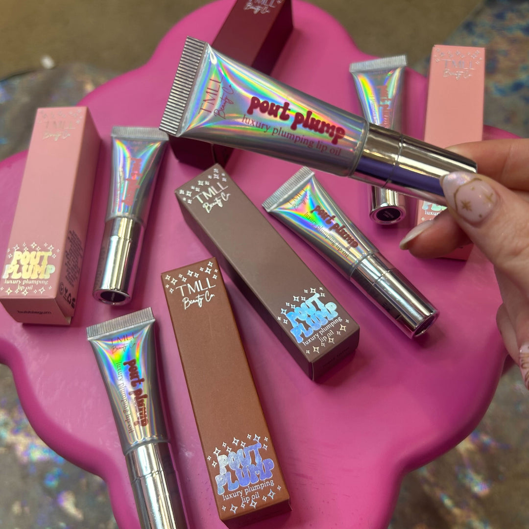 Hand reaching for TMLL Beauty Co Pout Plump luxury plumping lip oil holographic silver tubes and boxes in pink and brown shades arranged on pink surface