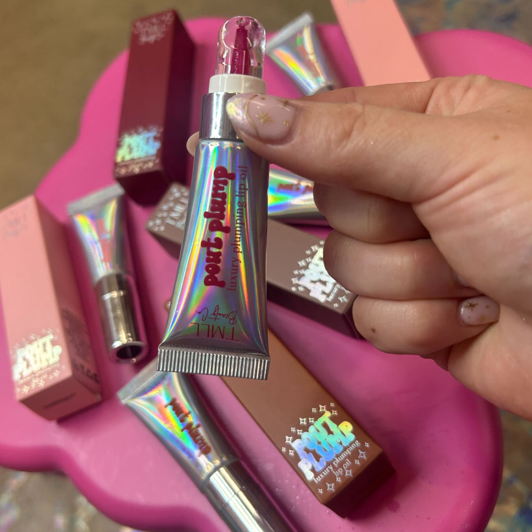 Hand holding TMLL Beauty Co Pout Plump luxury plumping lip oil holographic silver tube with pink glitter applicator cap, surrounded by additional tubes and boxes on pink surface