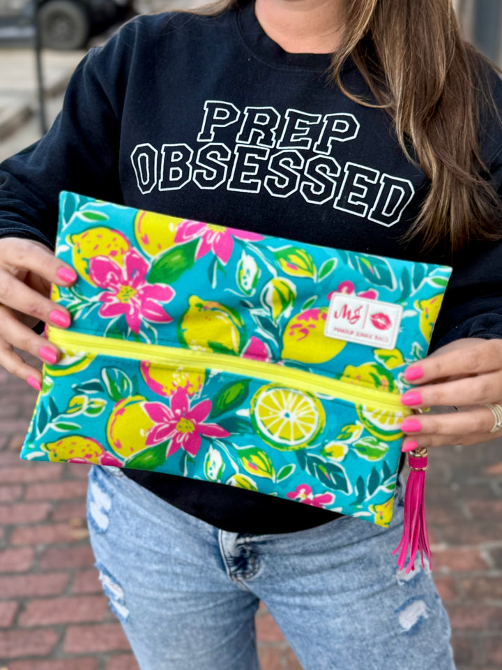 Model holding Makeup Junkie Bags large fabric zip clutch in teal lemon and floral print with yellow zipper and pink tassel while wearing Prep Obsessed sweatshirt outdoors