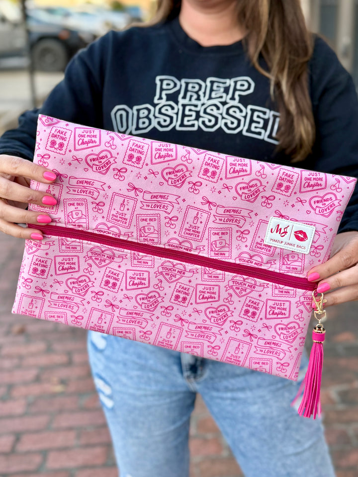 Model holding Makeup Junkie Bags large zip clutch in pink bookish romance print with hot pink glitter zipper and pink tassel while wearing Prep Obsessed sweatshirt outdoors