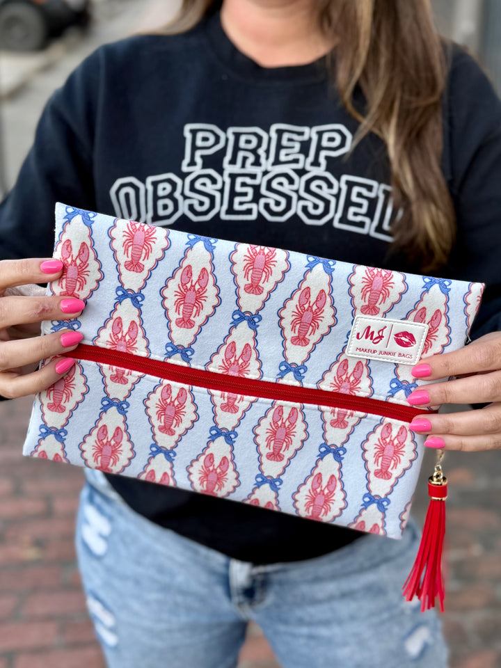 Model holding Makeup Junkie Bags large flat zip clutch in pink and blue lobster print with red zipper and red tassel while wearing Prep Obsessed sweatshirt outdoors