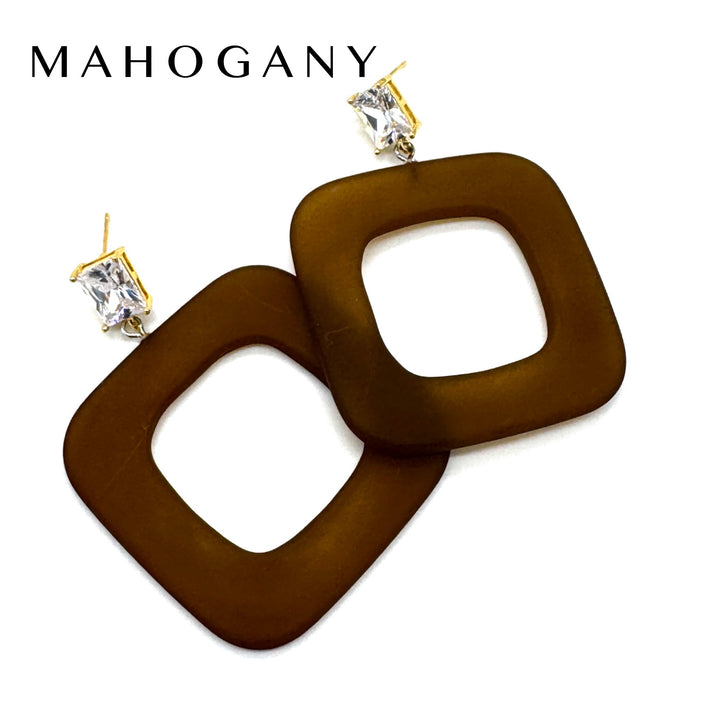 A pair of Mahogany statement earrings featuring large, square-shaped brown translucent discs with gold post and clear rhinestone studs, shown on a white background with the brand name 'MAHOGANY' printed at the top.
