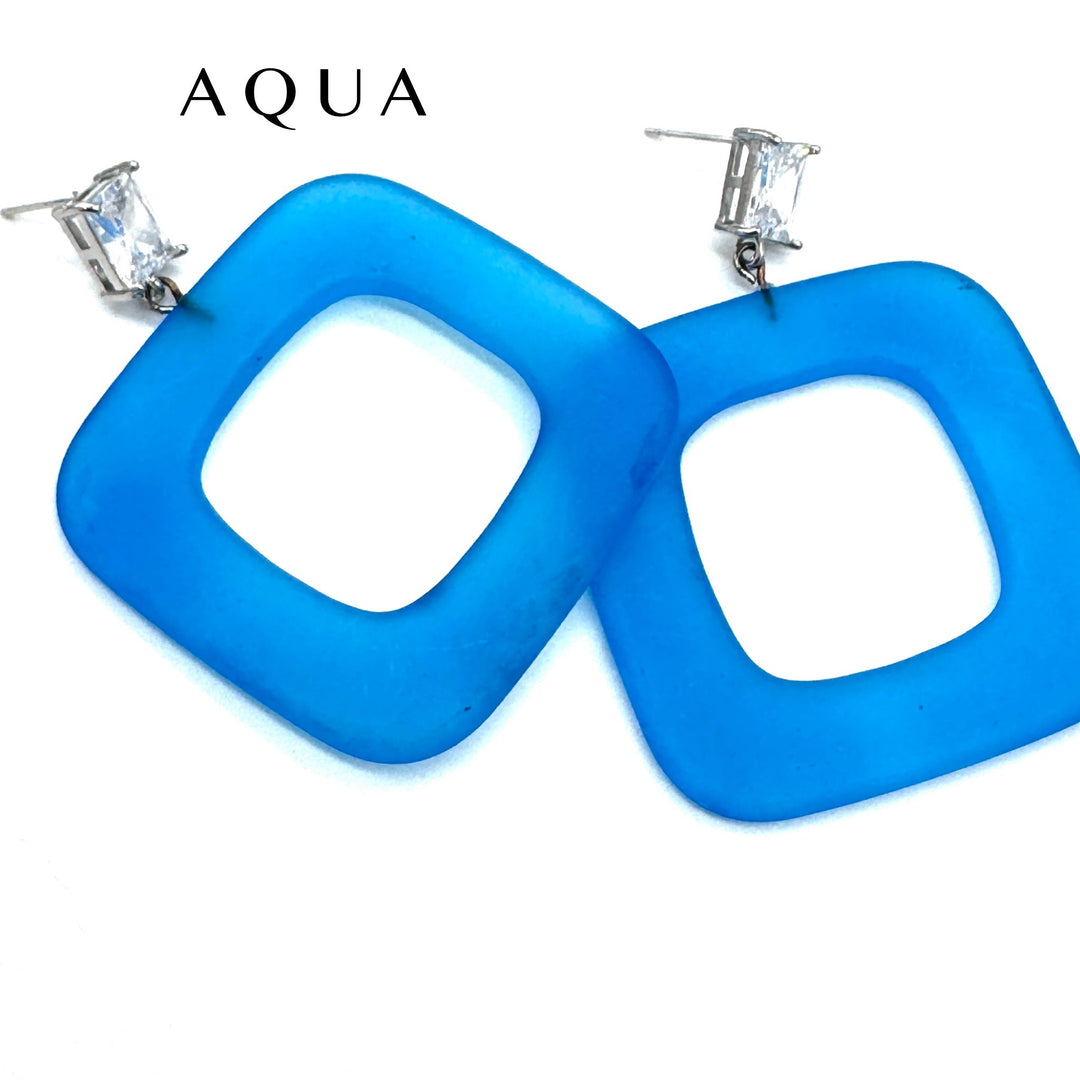 A pair of large square-shaped aqua blue lucite earrings with clear rectangular rhinestones on silver posts, shown against a plain white background with the brand name 'AQUA' printed above.