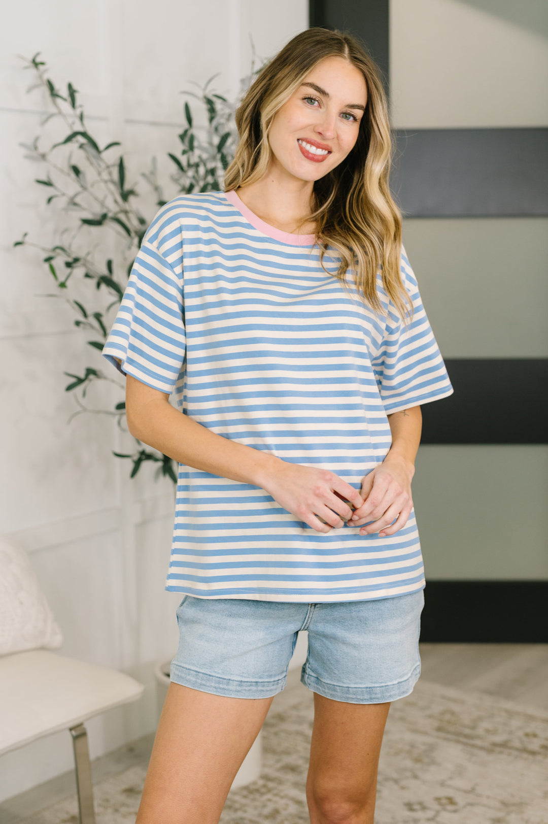 Woman wearing a blue and white striped short-sleeve top with denim shorts, posed in front of a white bench and leafy plant, full-length view