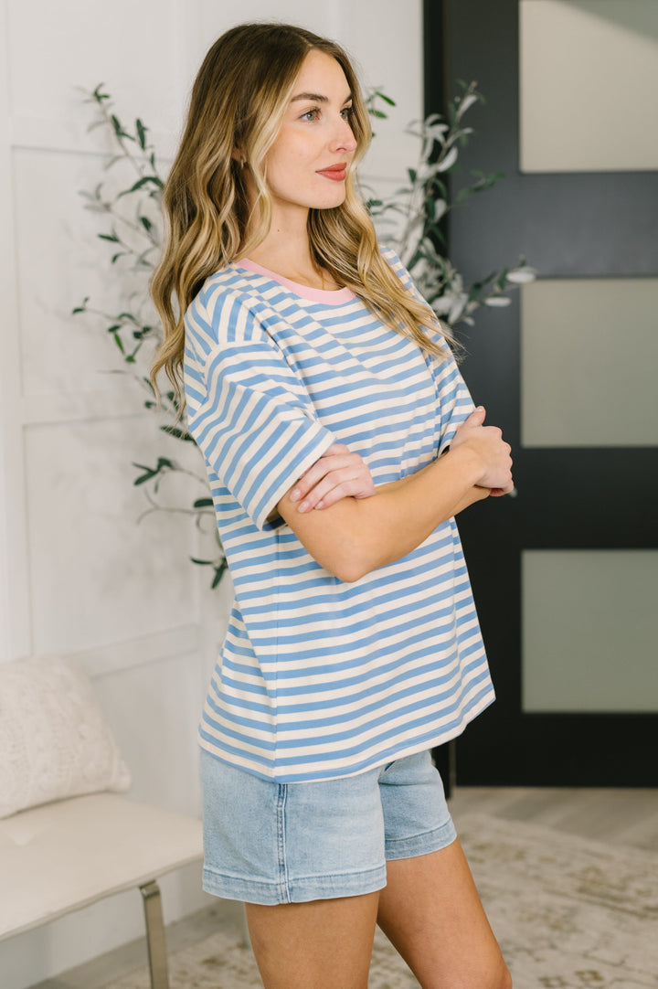 Side view of a woman wearing a blue and white striped short-sleeve top with denim shorts, arms crossed, posed in front of a white bench and leafy plant
