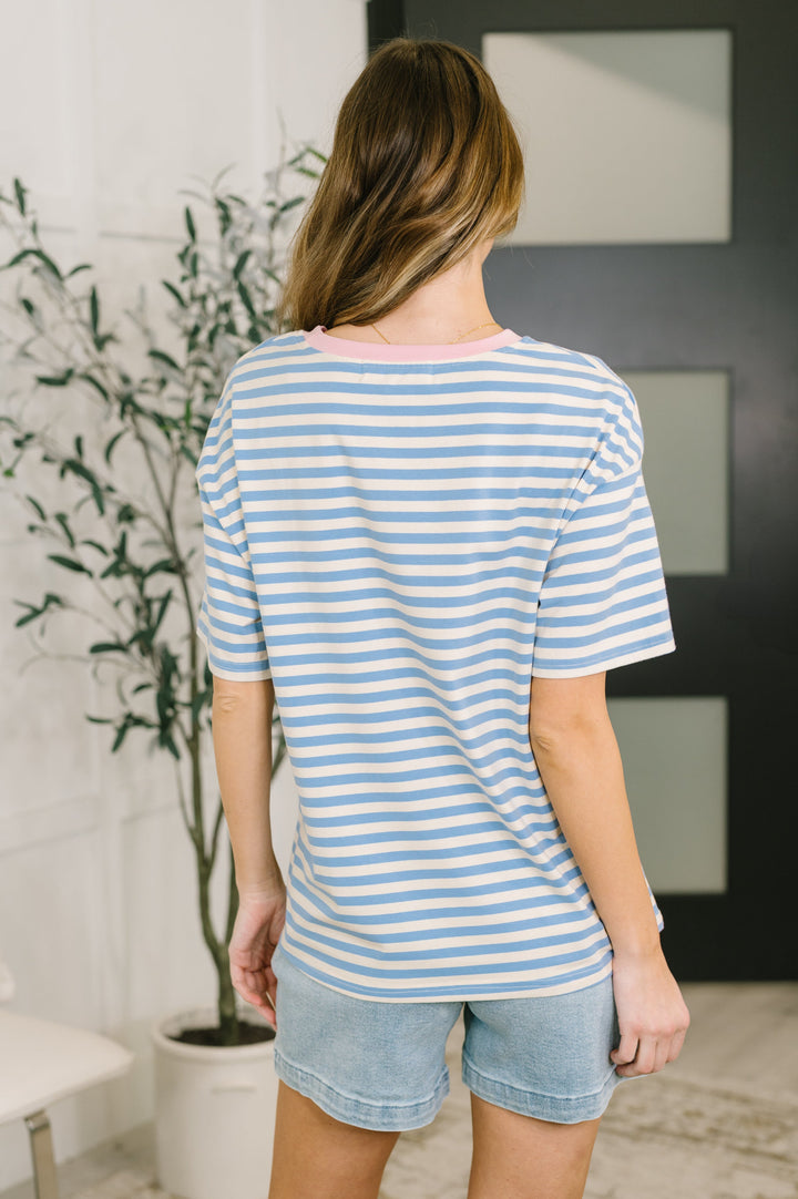 Back view of a woman wearing a blue and white striped short-sleeve top with denim shorts, arms crossed, posed in front of a white bench and leafy plant