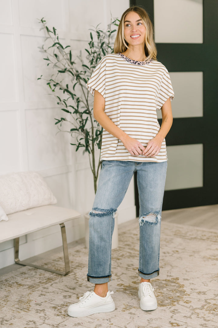 Woman wearing a white and tan striped oversized tee with pink leopard print neckline trim and side slits, styled with cuffed light wash jeans and white sneakers.