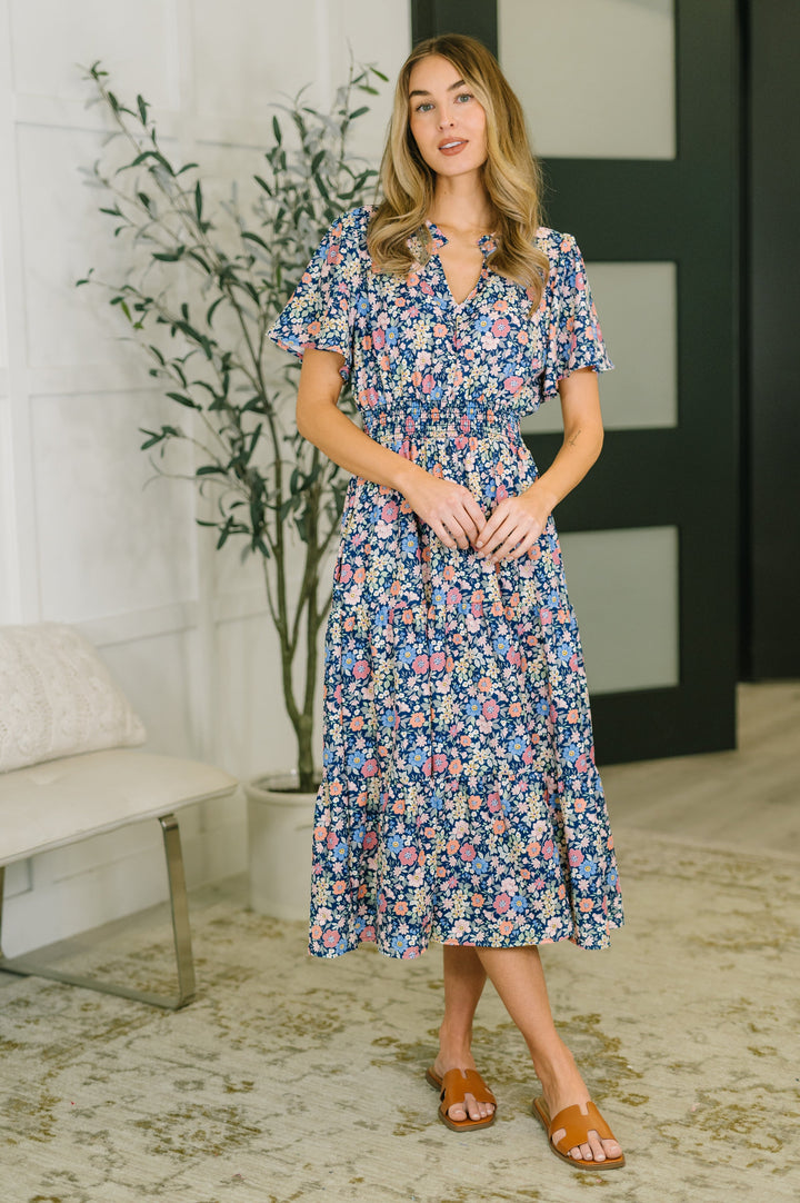 Woman wearing a navy ditsy floral midi dress with ruffle V-neckline, short flutter sleeves, smocked waist, and tiered skirt, styled with tan slide sandals.