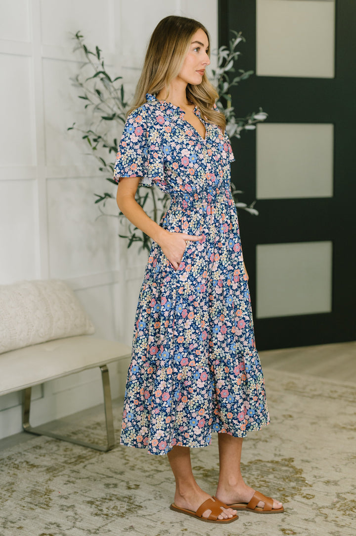 Side view of woman wearing a navy ditsy floral midi dress with smocked waist and side pocket, styled with tan slide sandals.