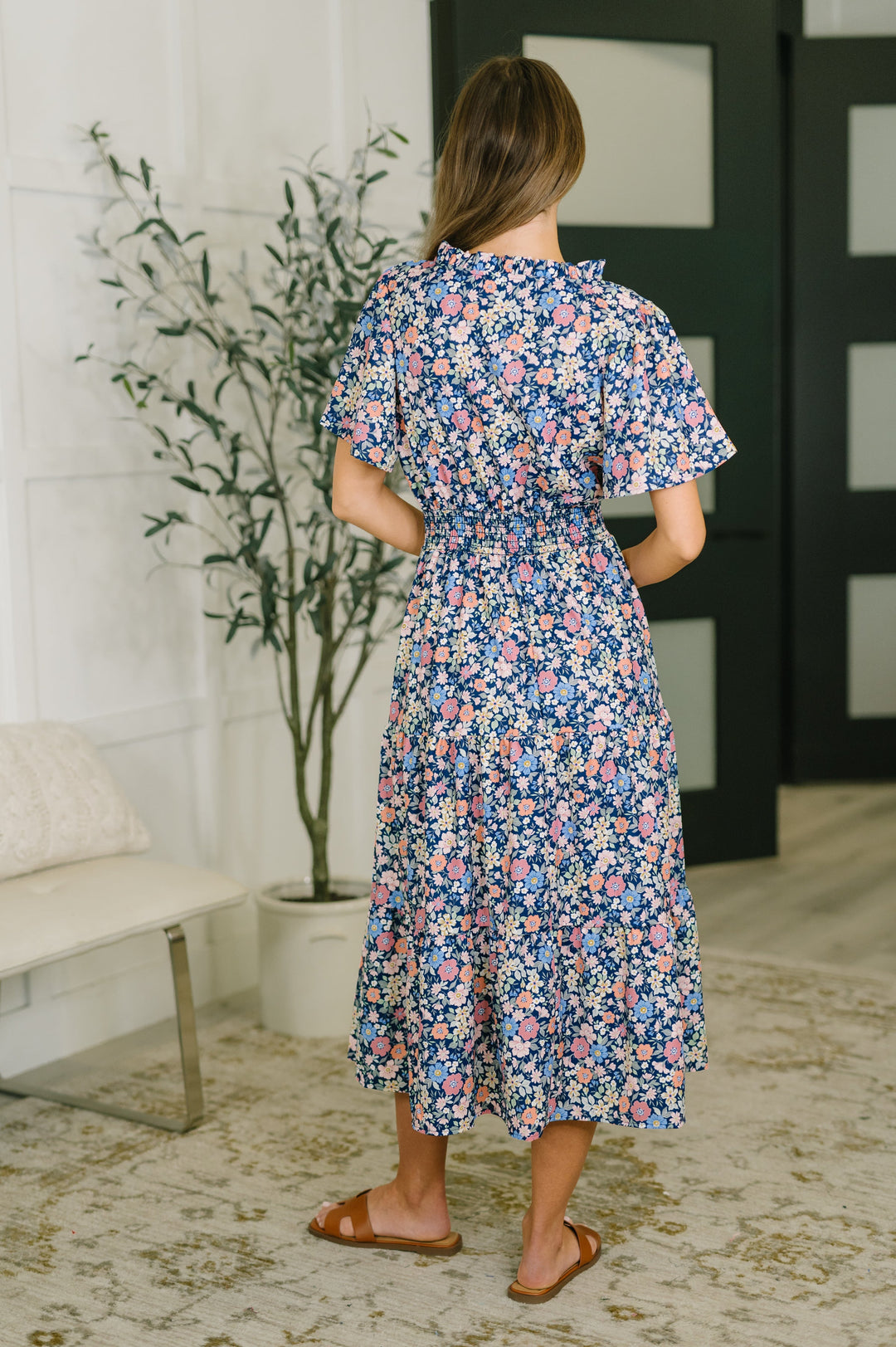 Back view of navy ditsy floral midi dress showing flutter sleeves, smocked waist, and tiered midi skirt, styled with tan slide sandals.