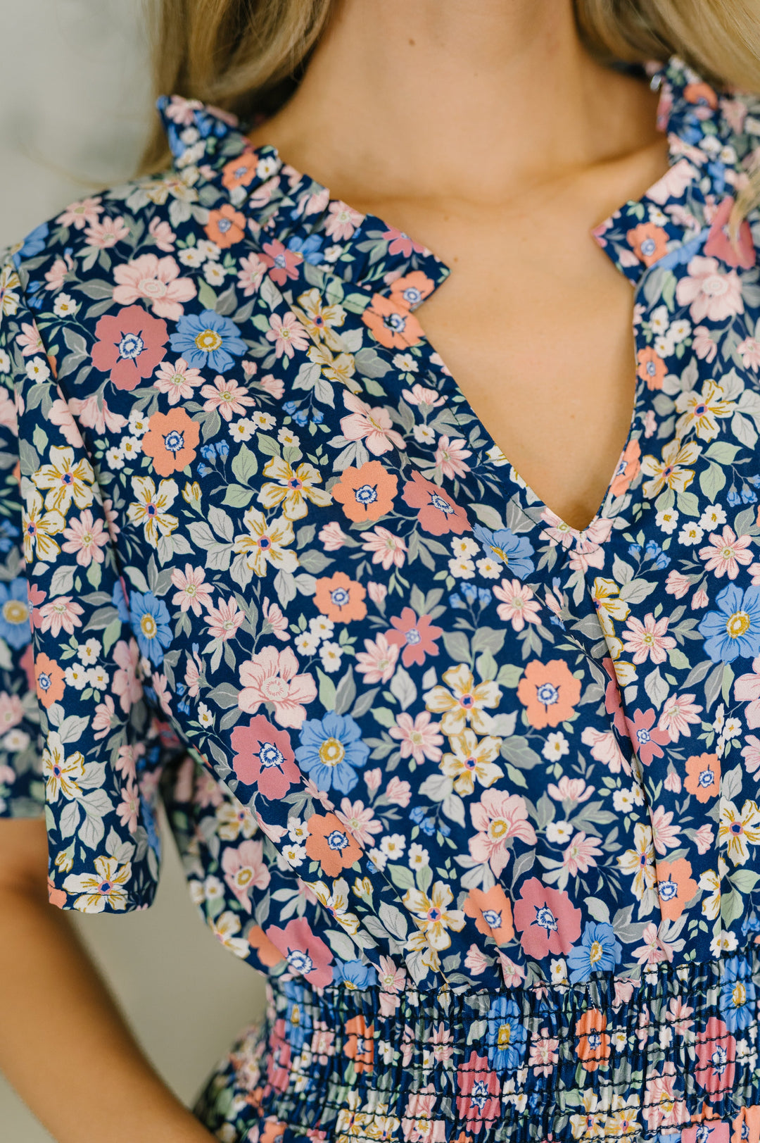 Close-up of navy ditsy floral midi dress showing ruffle V-neckline, pink and blue floral print, and smocked waist detail.
