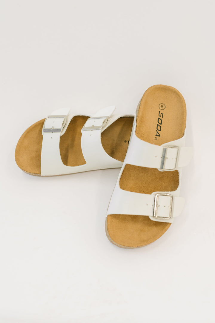 Angled side view of white double-buckle slide sandals by Soda with a tan cork footbed, on a white background.